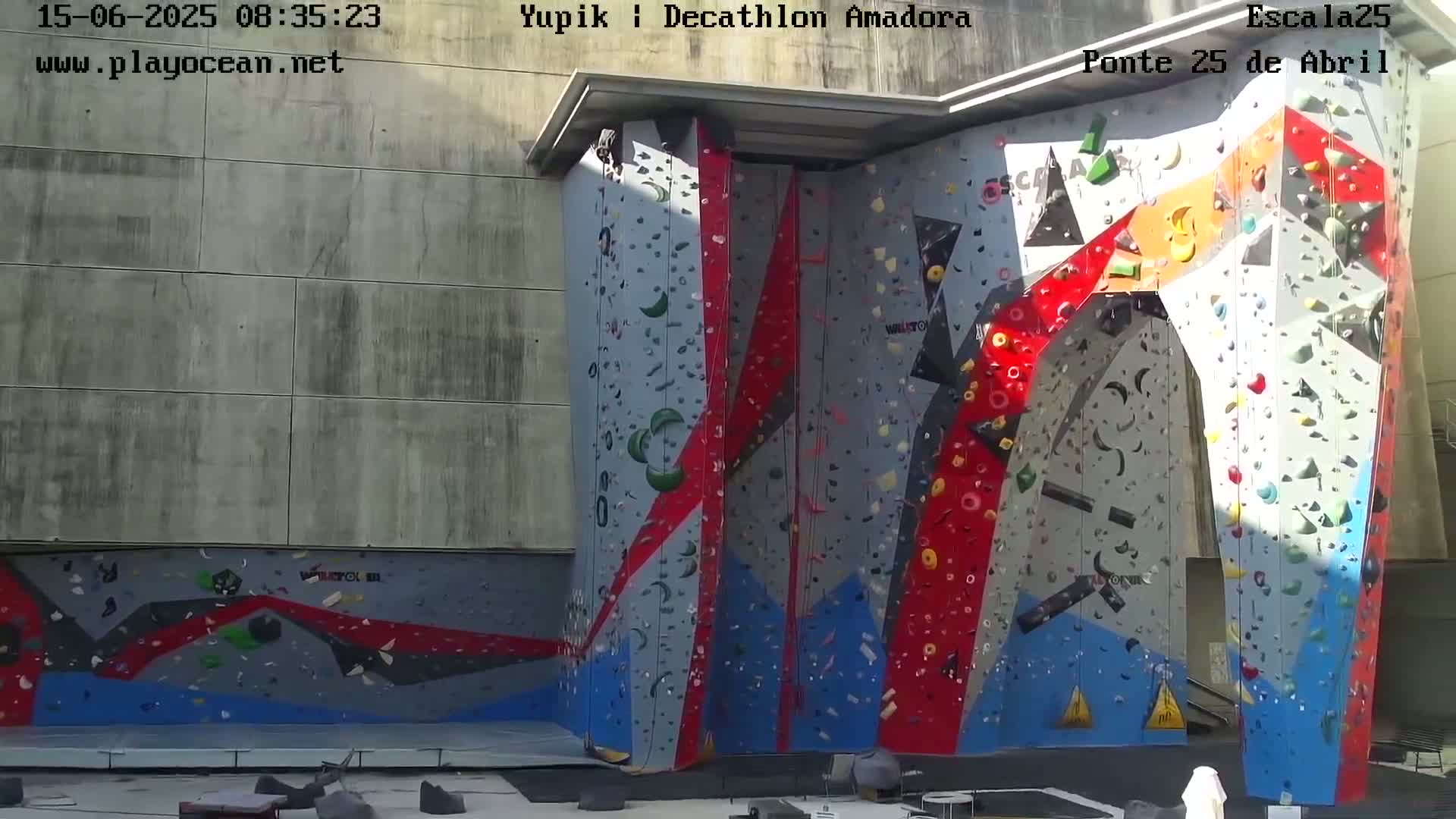 A large, multi-colored climbing wall stands outdoors in bright sunlight.