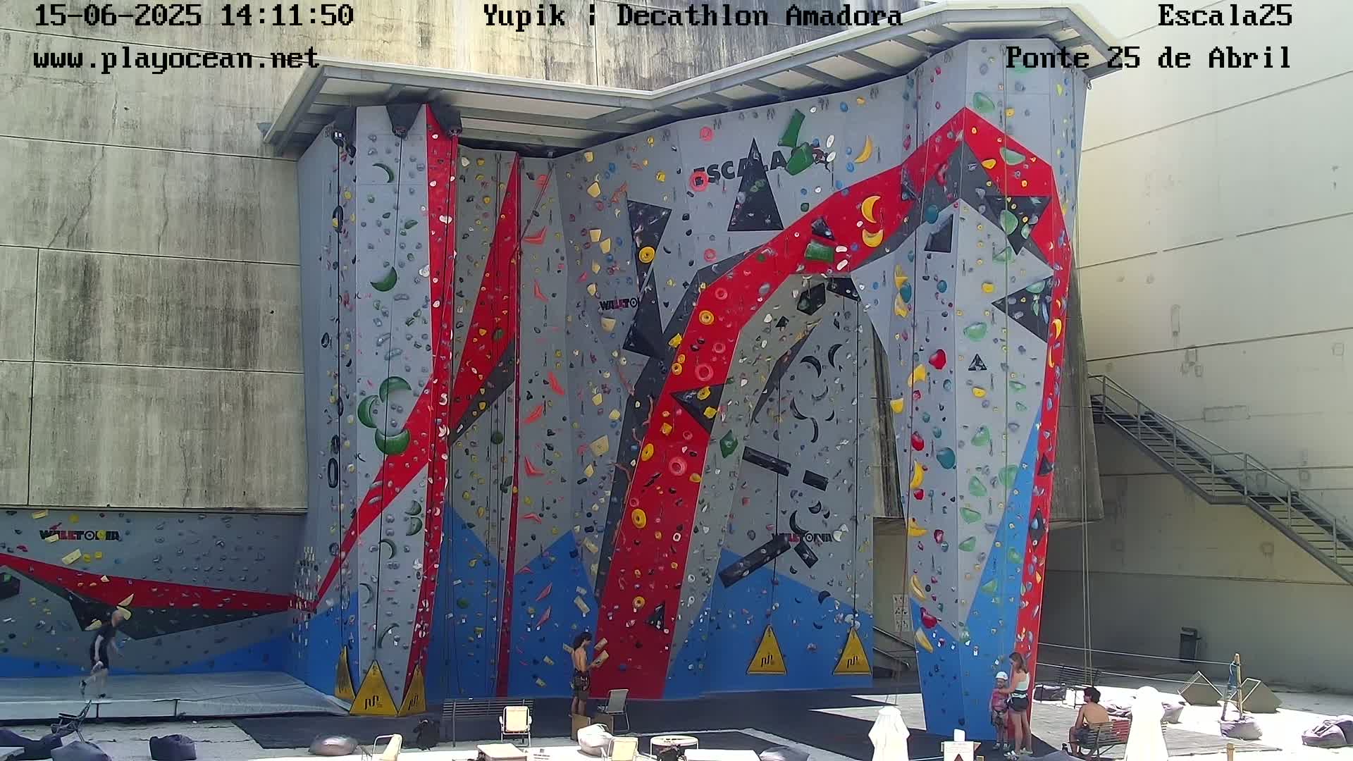 A large, colorful outdoor climbing wall with several people near it on a sunny day.