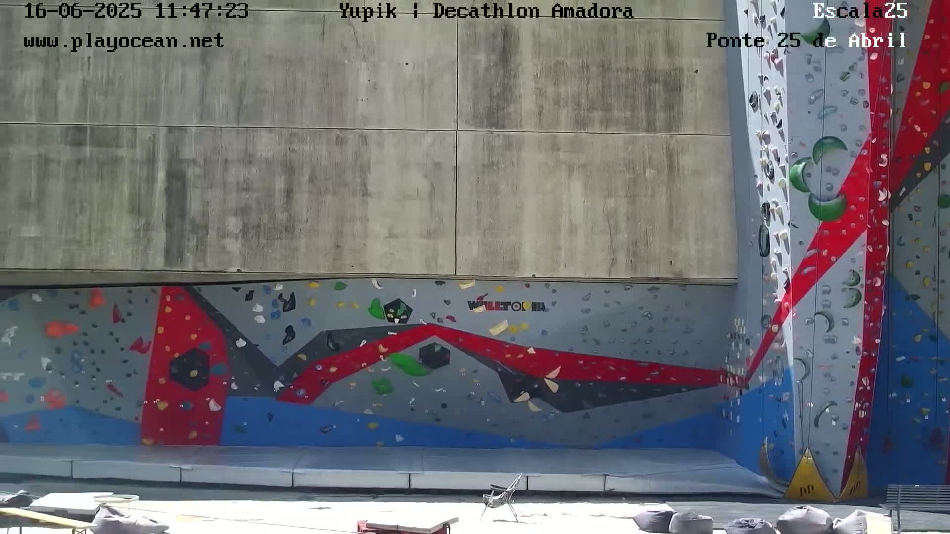 A large outdoor climbing wall with colorful geometric designs is situated against a concrete wall on a sunny day.