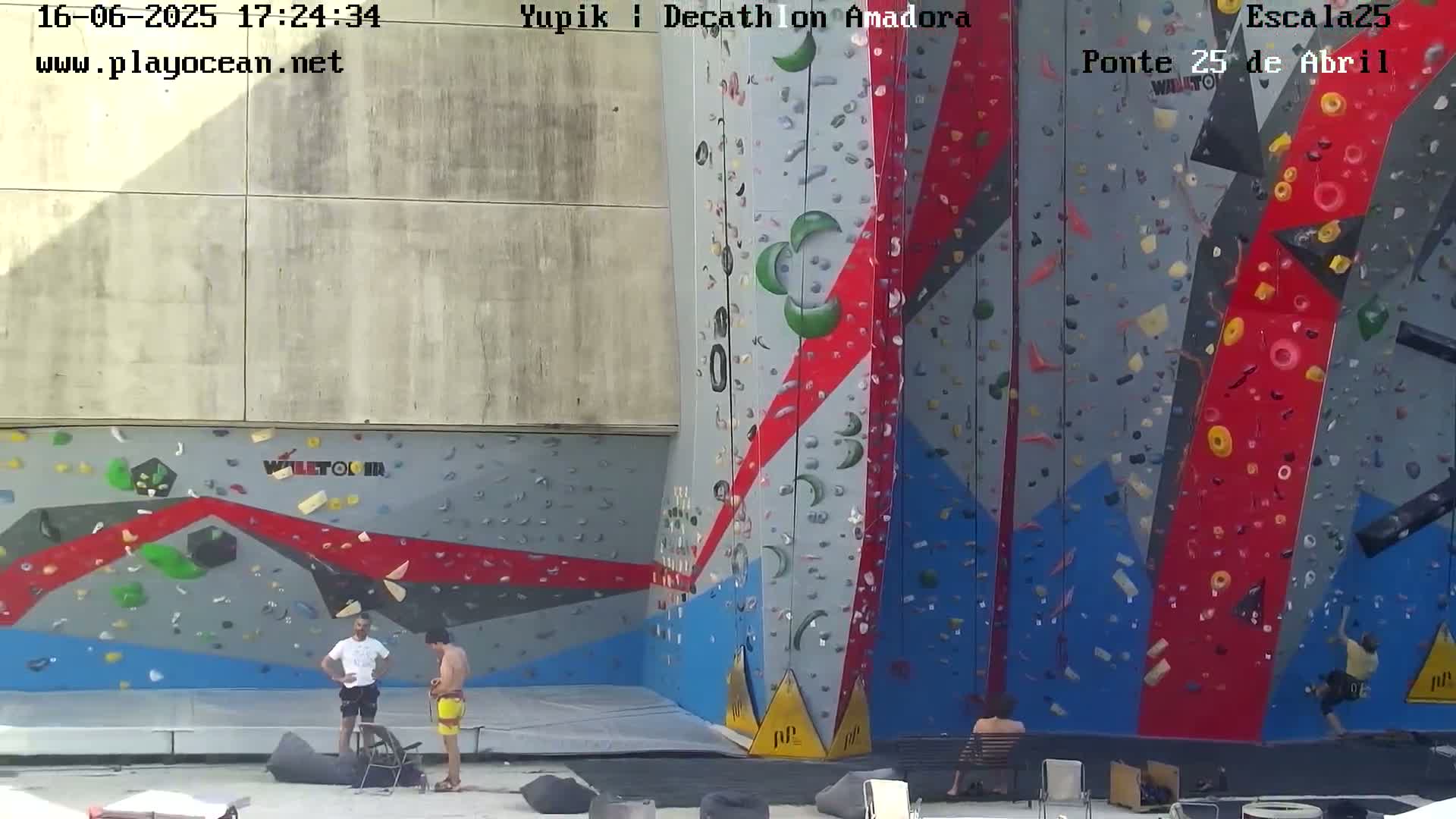 Two men stand near a large, colorful outdoor climbing wall on a sunny day.