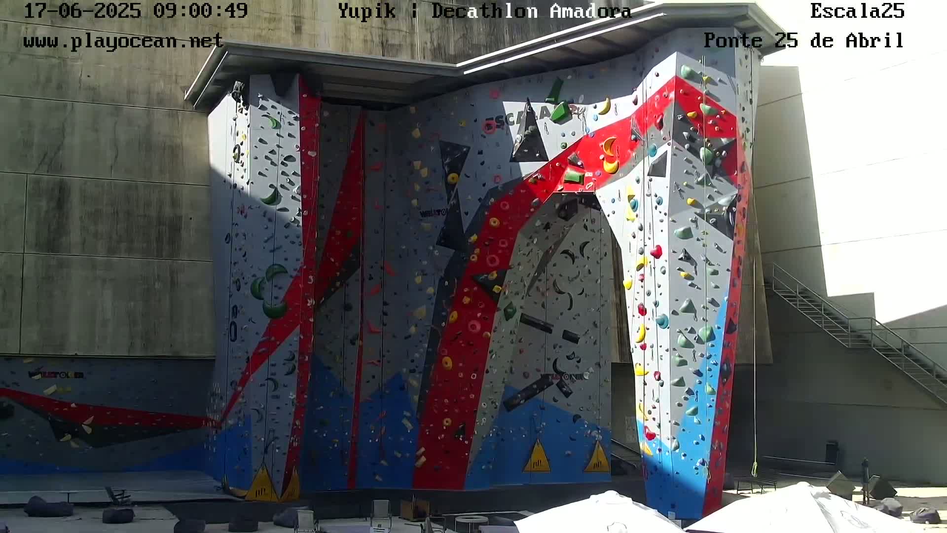 A large, colorful indoor climbing wall is situated outdoors in bright sunlight.