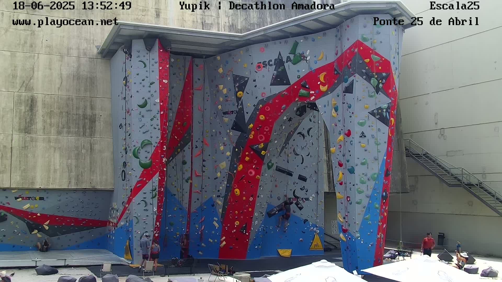 A large outdoor climbing wall with various colored holds, some people are at the base of the wall and a few people are climbing.