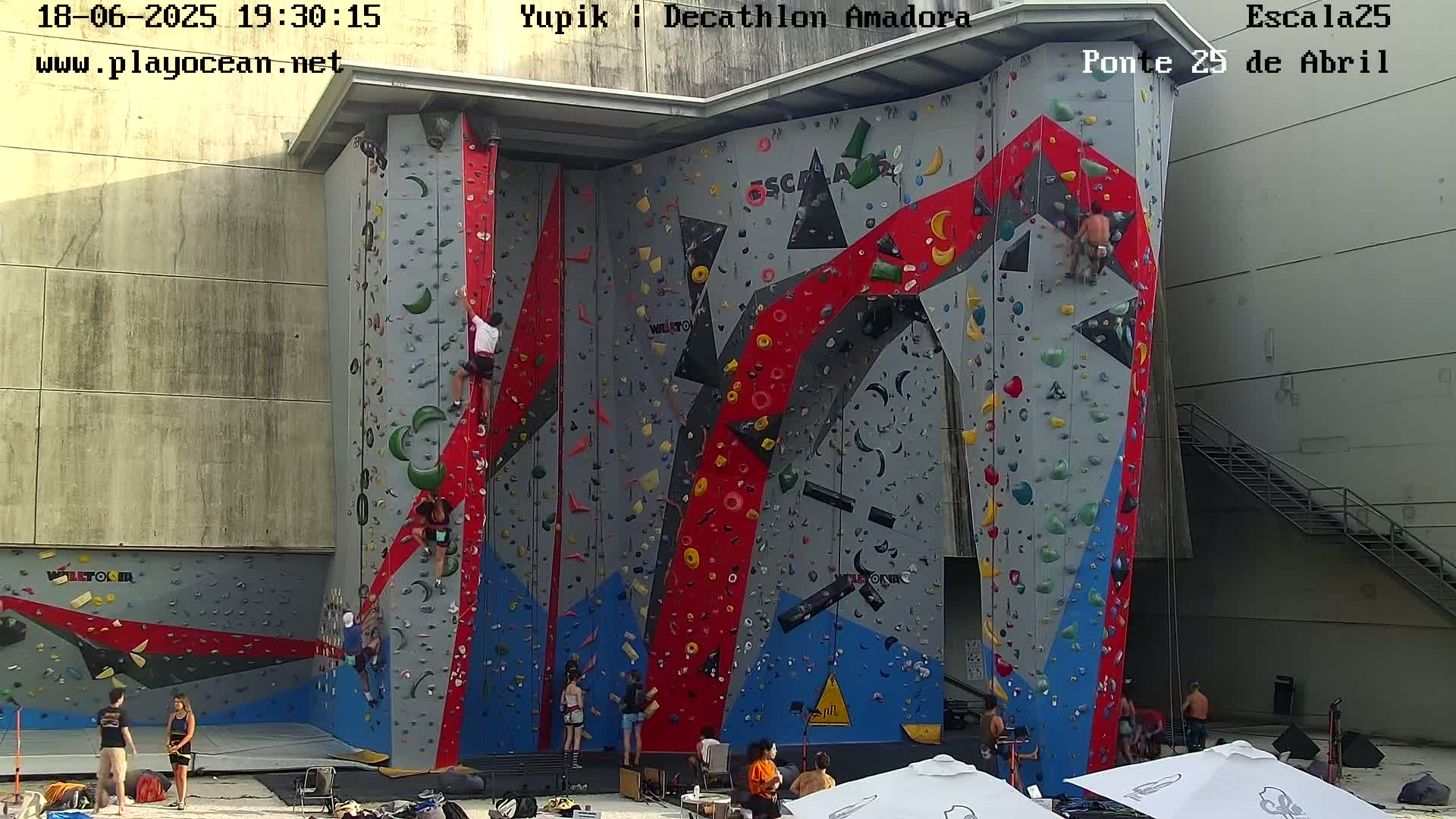 Several people are rock climbing on a large, colorful outdoor climbing wall on a sunny day.
