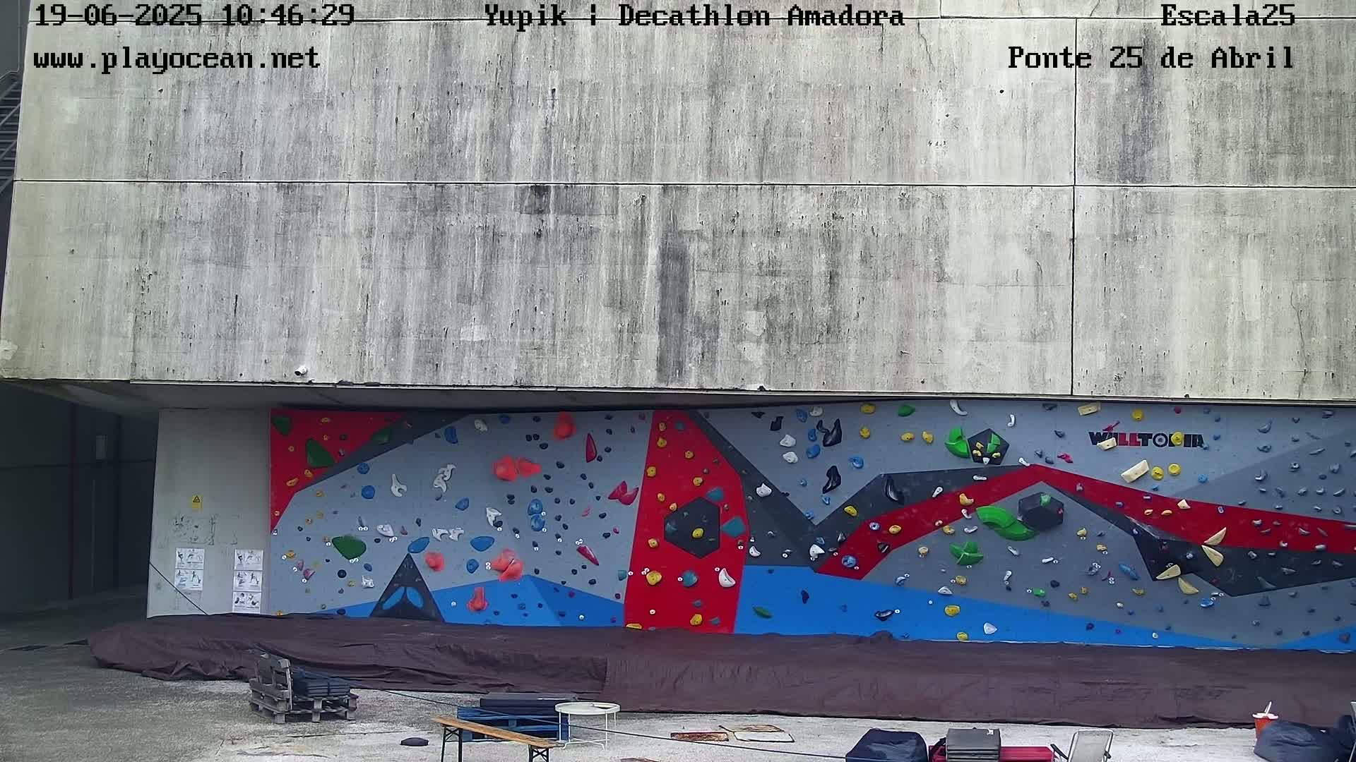 A large, colorful outdoor climbing wall is situated against a gray concrete building under an overcast sky.