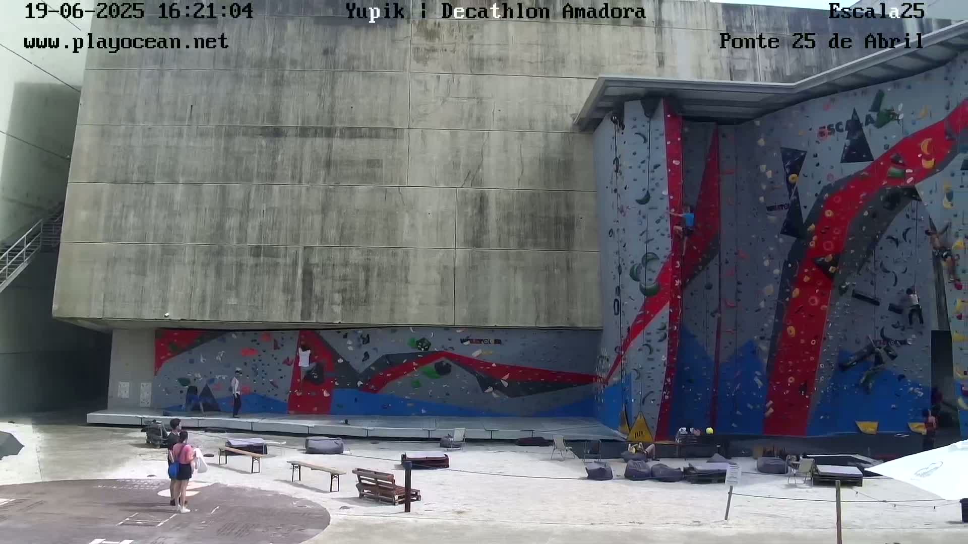 An outdoor climbing wall with several people climbing and others observing on a sunny day.
