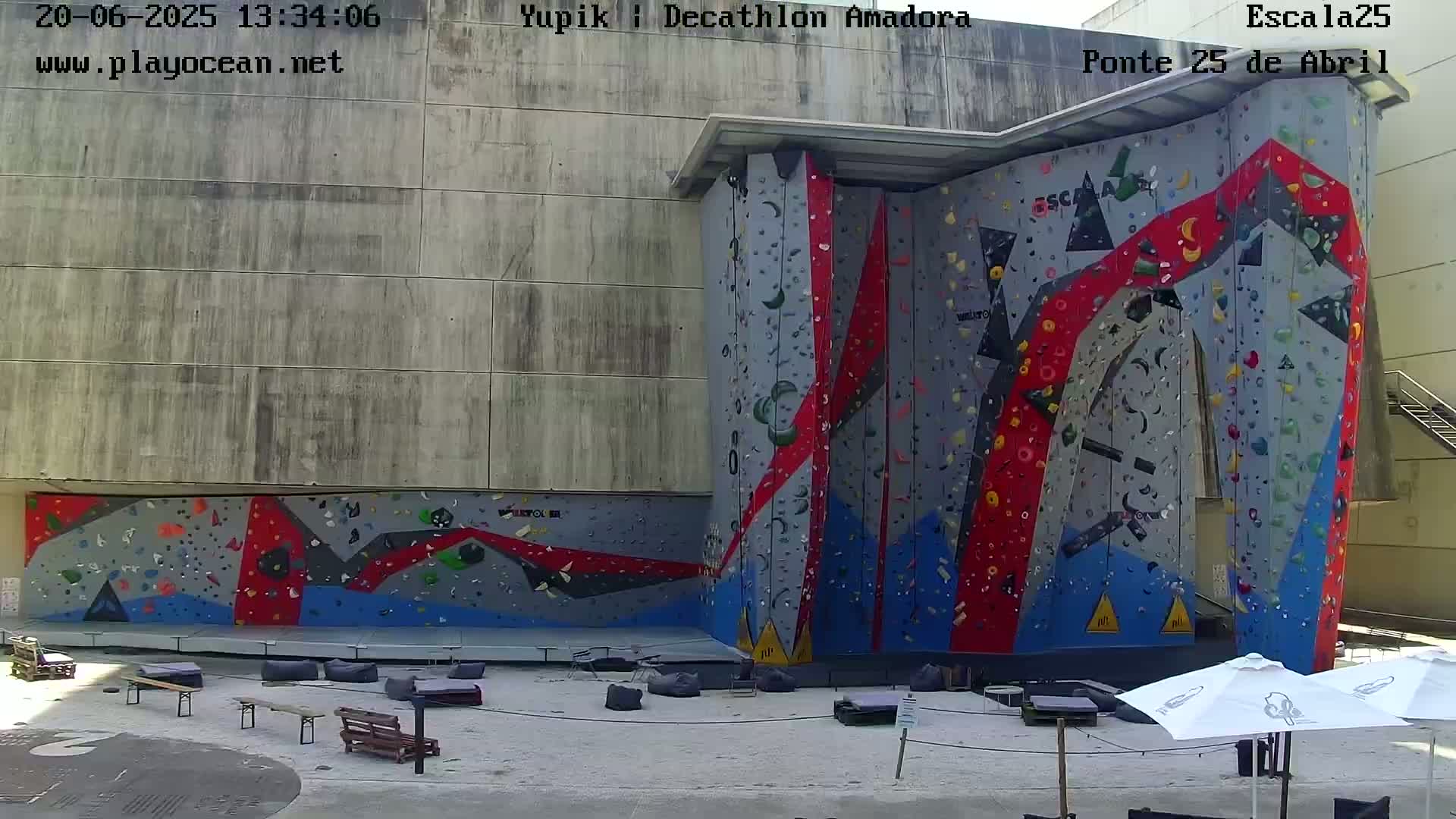 A large outdoor climbing wall with red, blue, and gray holds is situated against a gray building, with several benches and patio umbrellas in the foreground on a sunny day.