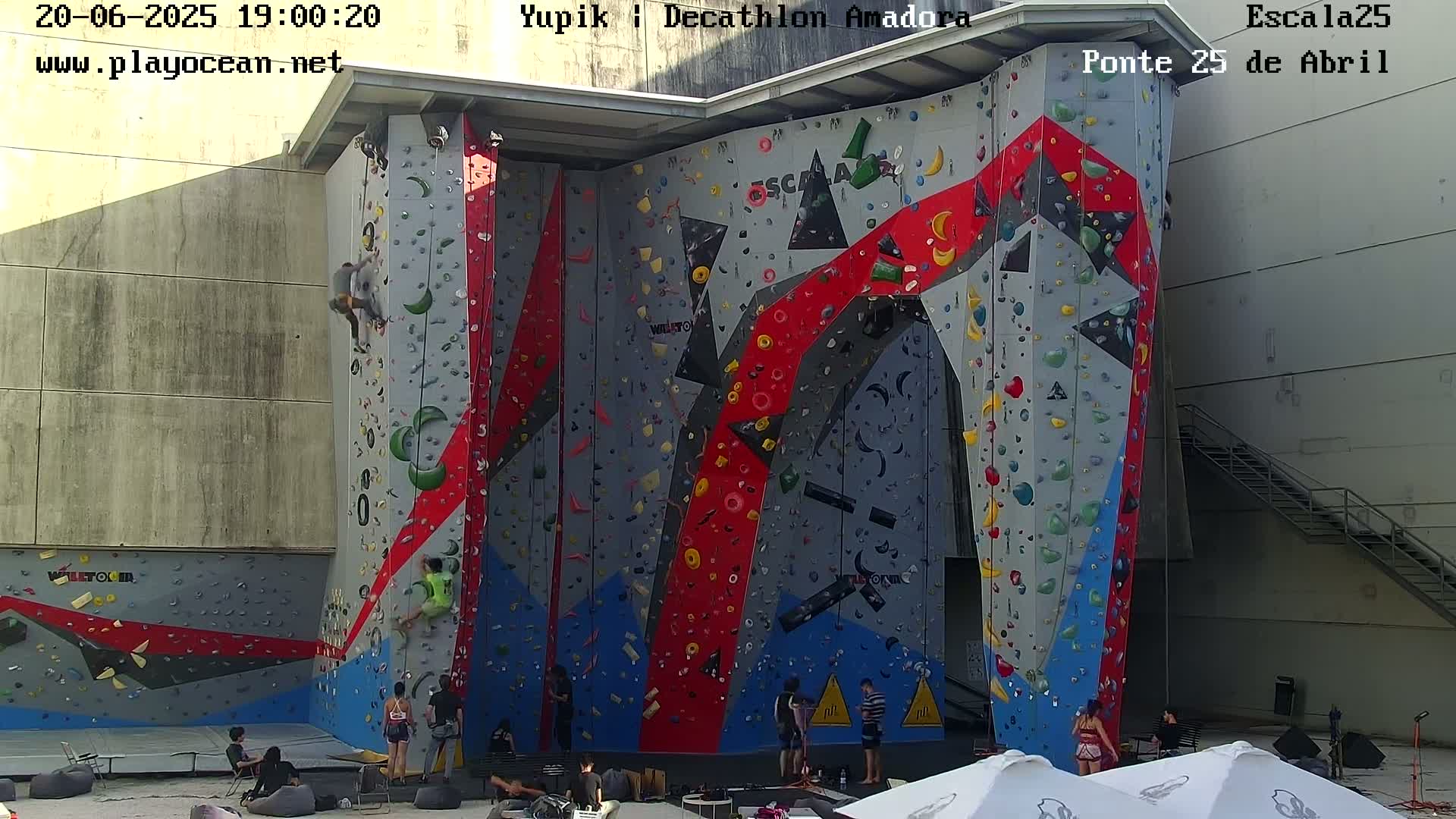 Several people are rock climbing on a large, multicolored indoor climbing wall, with others sitting and watching.
