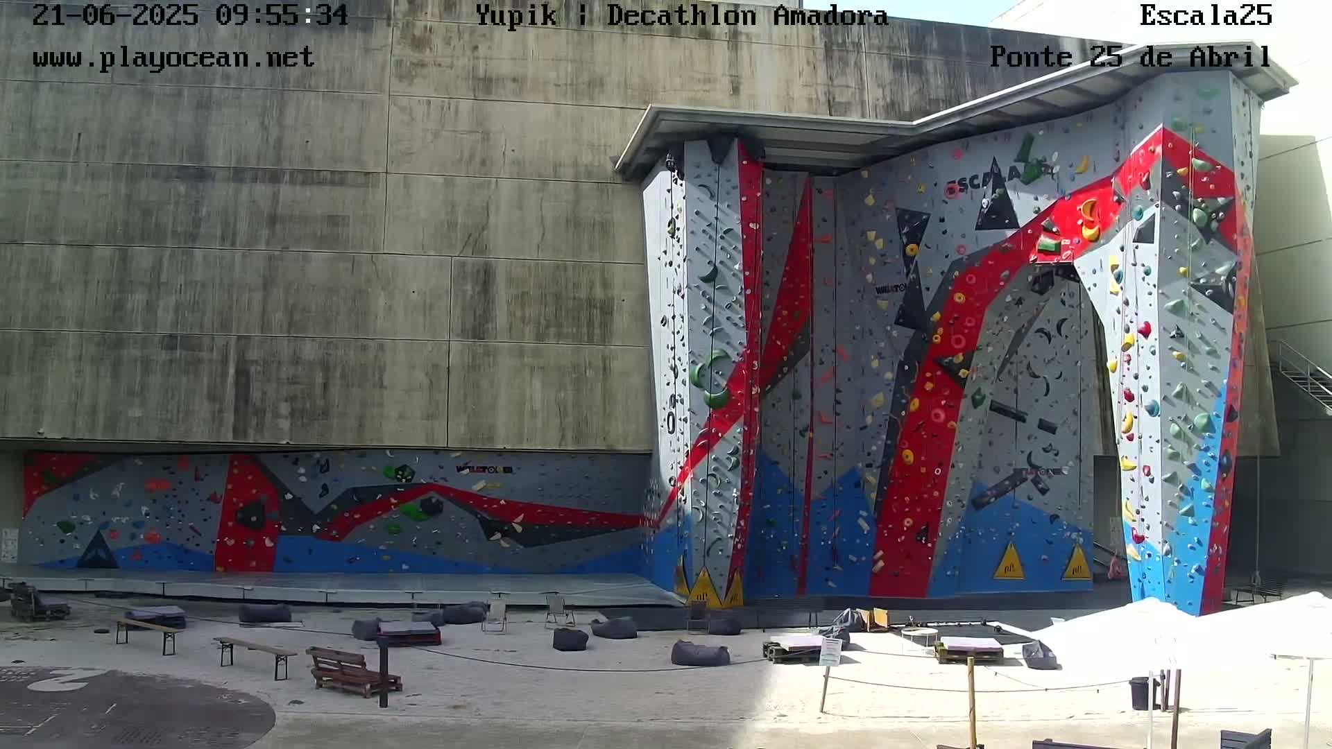 A large, colorful outdoor climbing wall is situated against a concrete building on a sunny day.