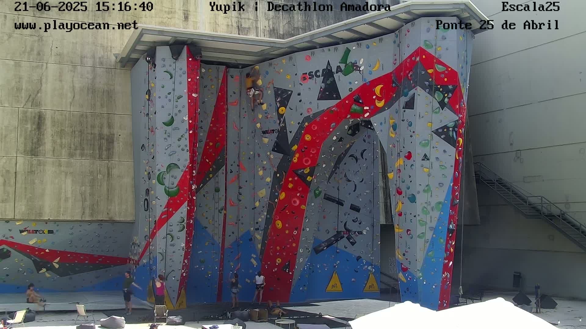 A large, colorful climbing wall is situated outdoors on a sunny day with several people climbing and watching at its base.