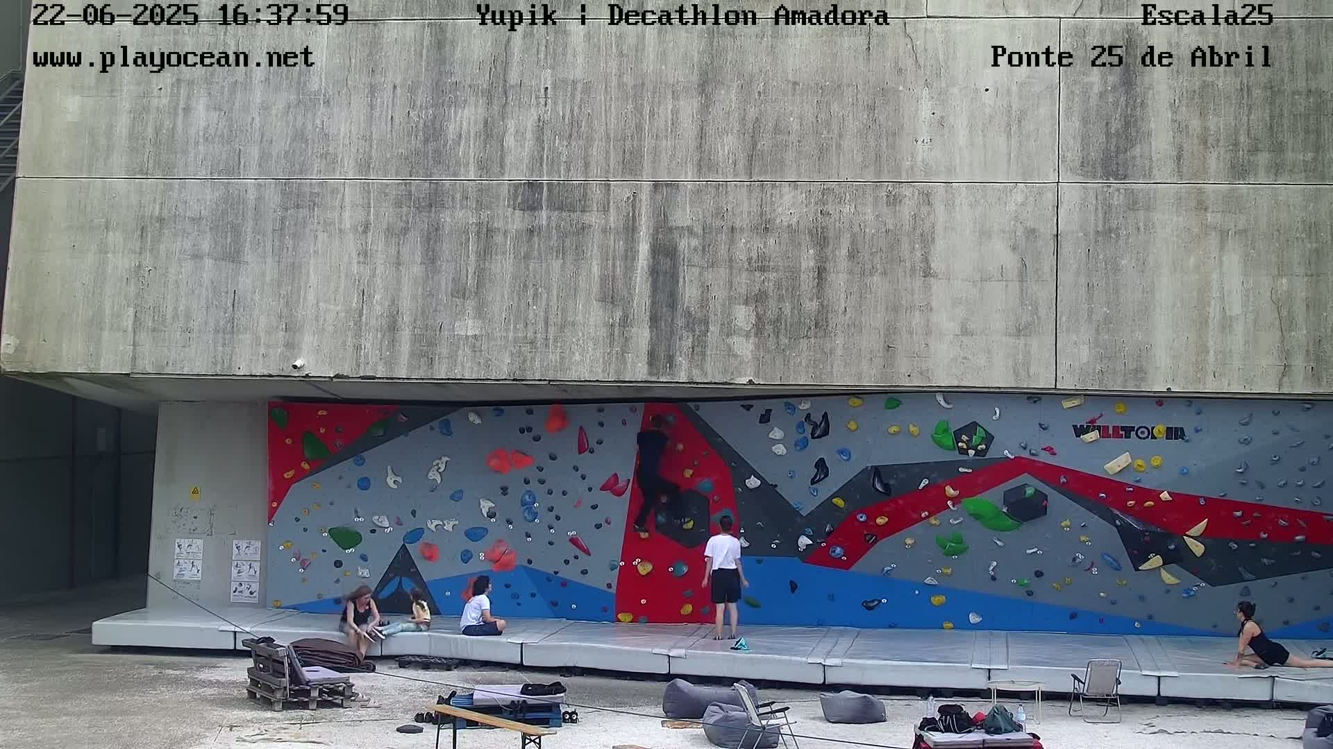 Several people are climbing and resting near a large, colorful outdoor climbing wall on a mostly cloudy day.
