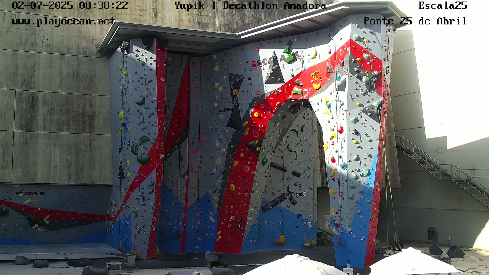 A large, colorful indoor climbing wall is situated outdoors under sunny conditions.