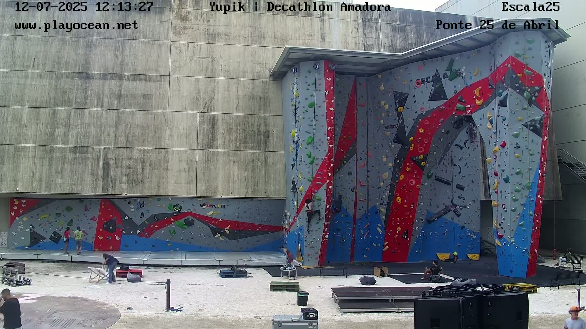 A large, colorful indoor climbing wall is situated outdoors under sunny conditions.