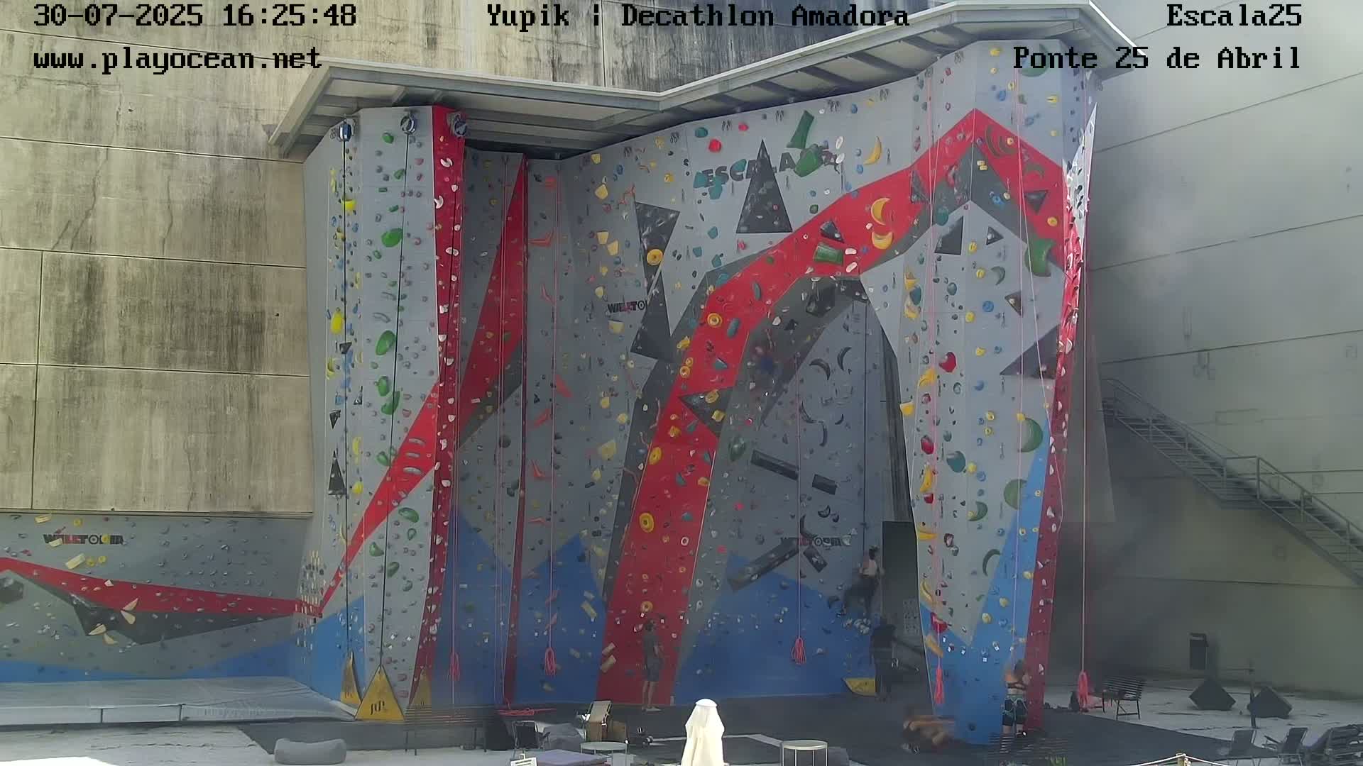 A colorful outdoor climbing wall is situated against a concrete building under overcast skies.