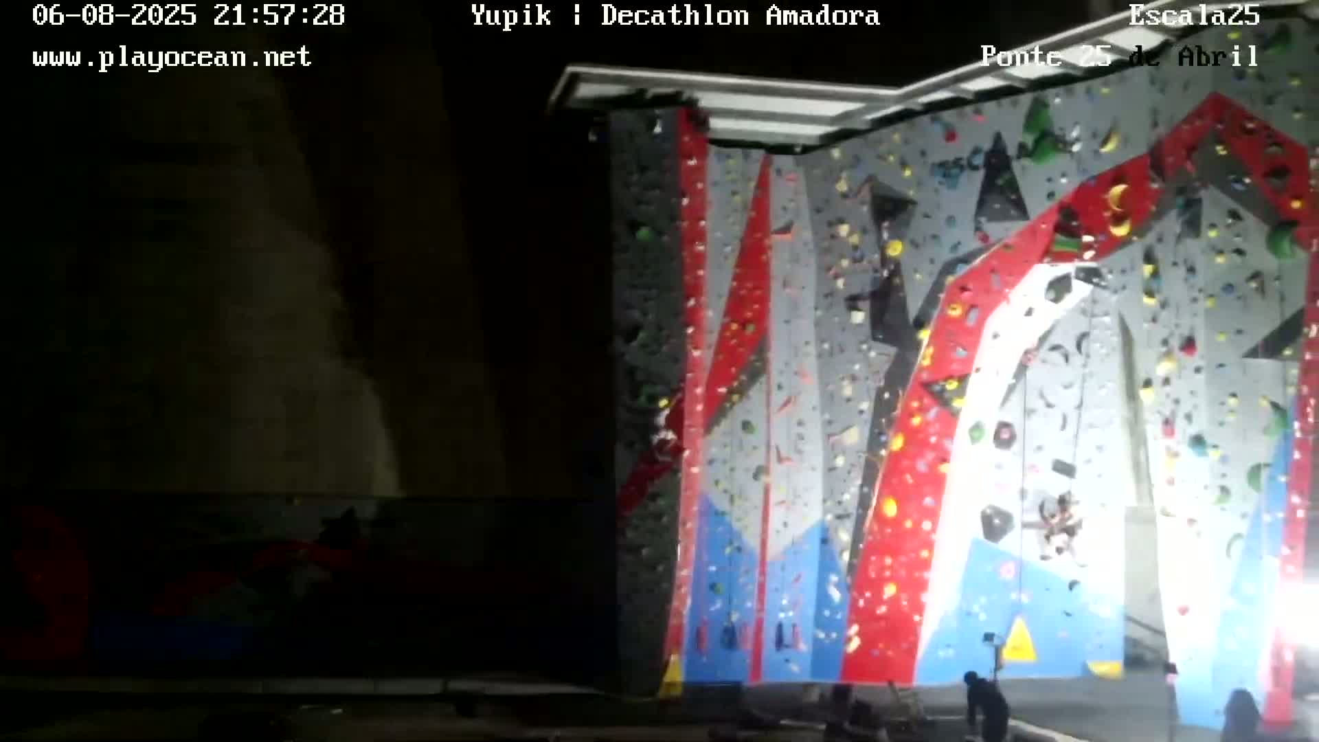 A large, brightly colored indoor climbing wall is shown at night, with a few people at its base.