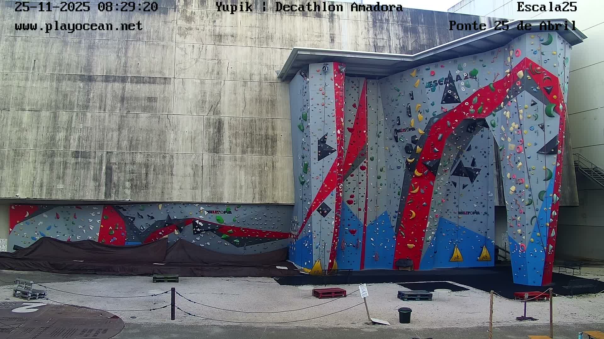 An expansive, grey, red, and blue outdoor climbing wall adorned with numerous colorful holds stands against a large concrete building under an overcast sky.