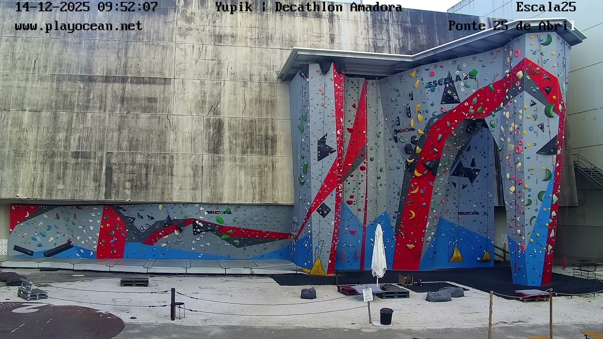 An expansive, grey, red, and blue outdoor climbing wall adorned with numerous colorful holds stands against a large concrete building under an overcast sky.