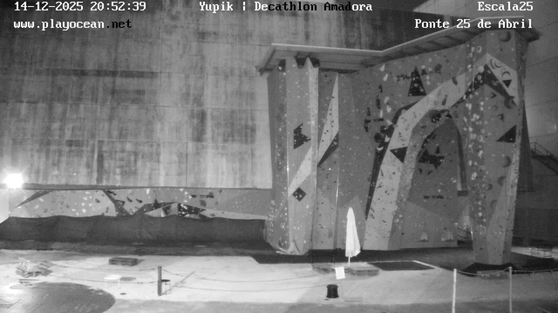 An expansive, grey, red, and blue outdoor climbing wall adorned with numerous colorful holds stands against a large concrete building under an overcast sky.