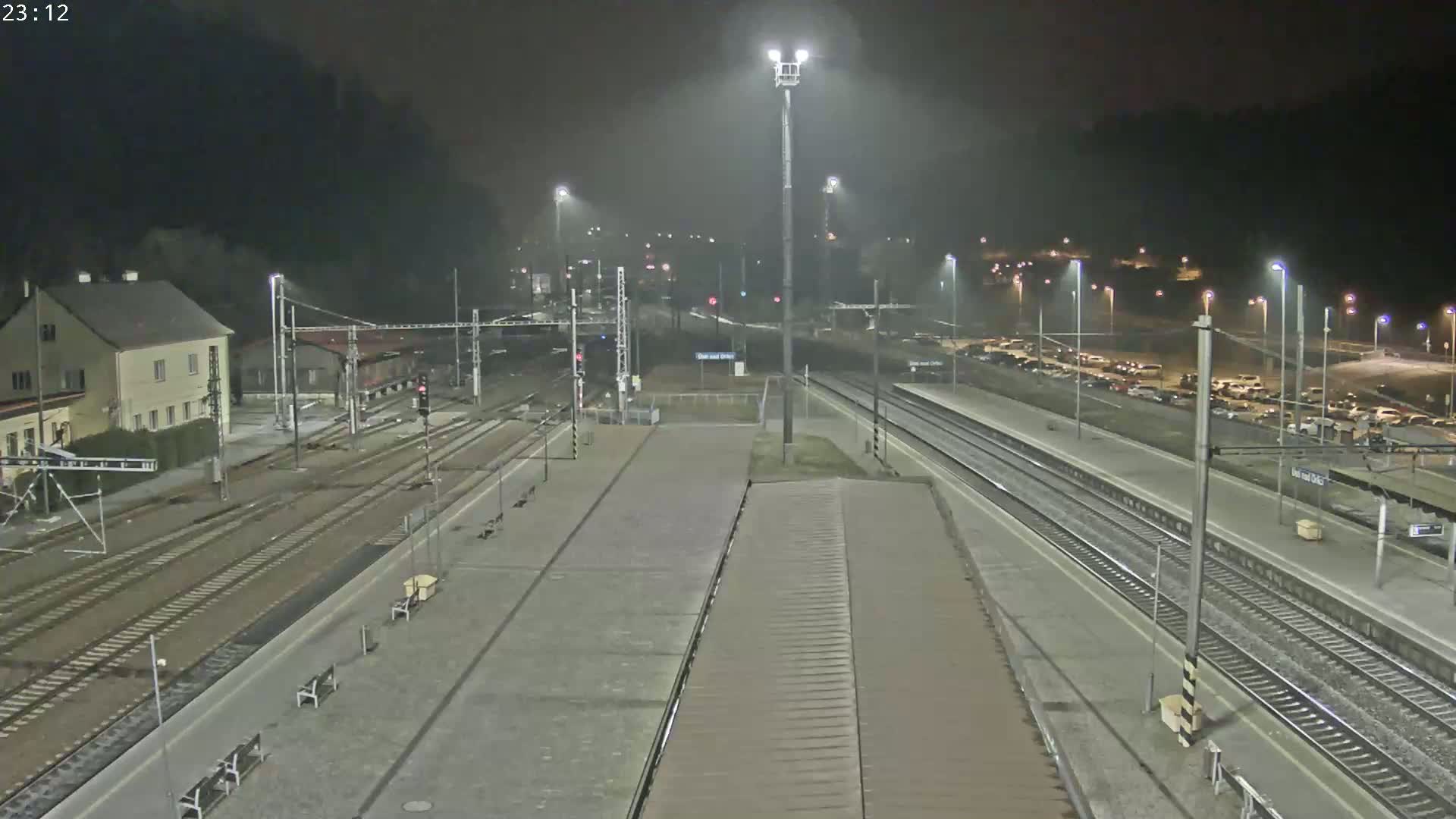A nighttime view of a train station with multiple tracks, a parking lot filled with cars, and a building in the background under hazy or foggy conditions.