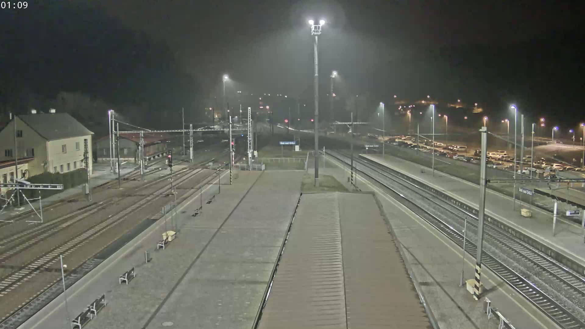 A nighttime view of a train station with multiple tracks, a building, and a parking lot in foggy conditions.