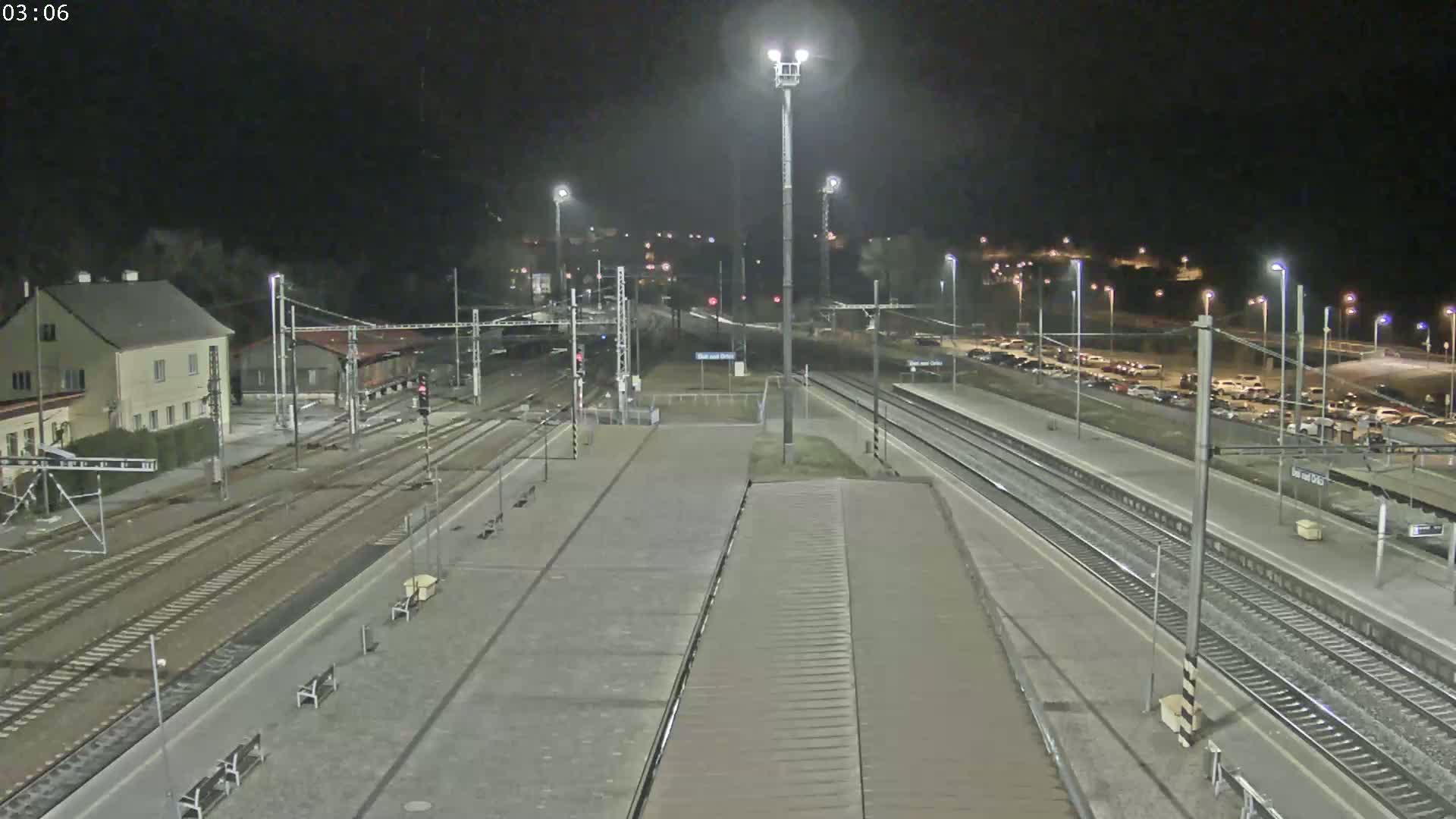 A nighttime view of a train station with multiple tracks, a platform, and a parking lot filled with cars under clear skies.