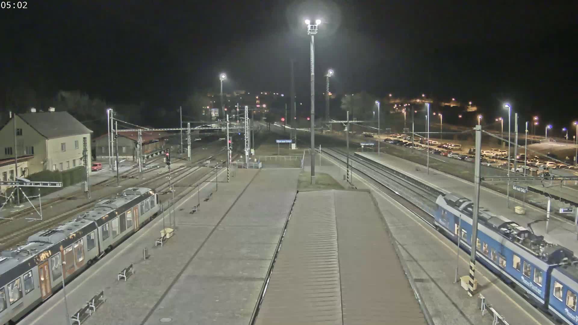 A nighttime view of a train station with two trains at the platform, numerous lights illuminating the scene, and cars parked in a lot.