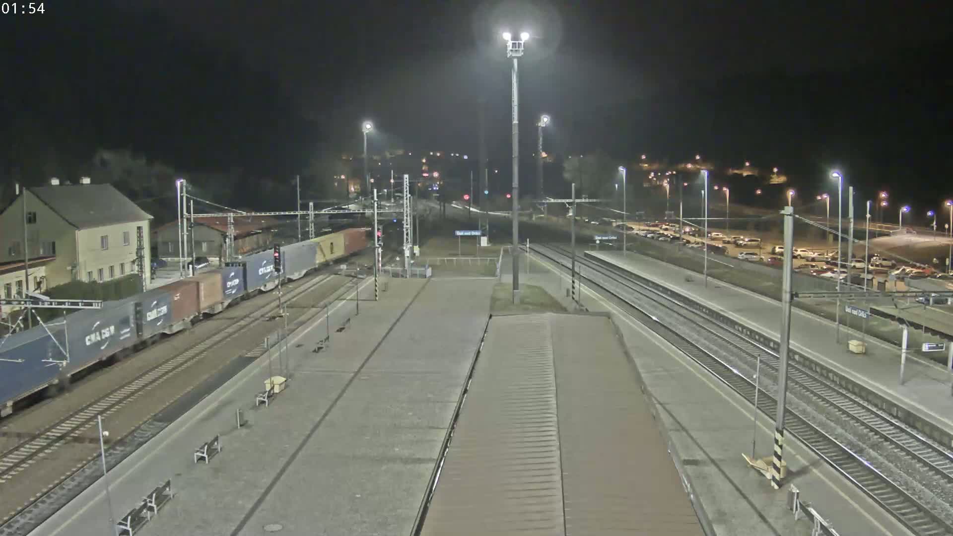 A nighttime view of a train station with a freight train on the tracks and a parking lot filled with cars.