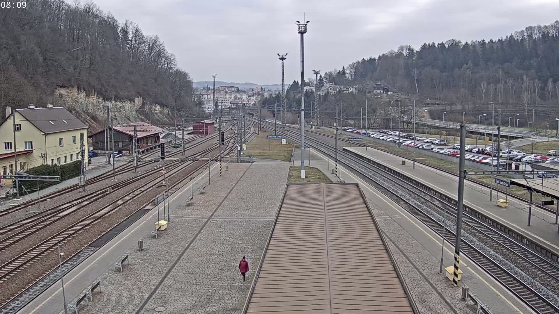 A train station with multiple tracks and a paved platform under an overcast sky, a person in the distance walking across the platform.