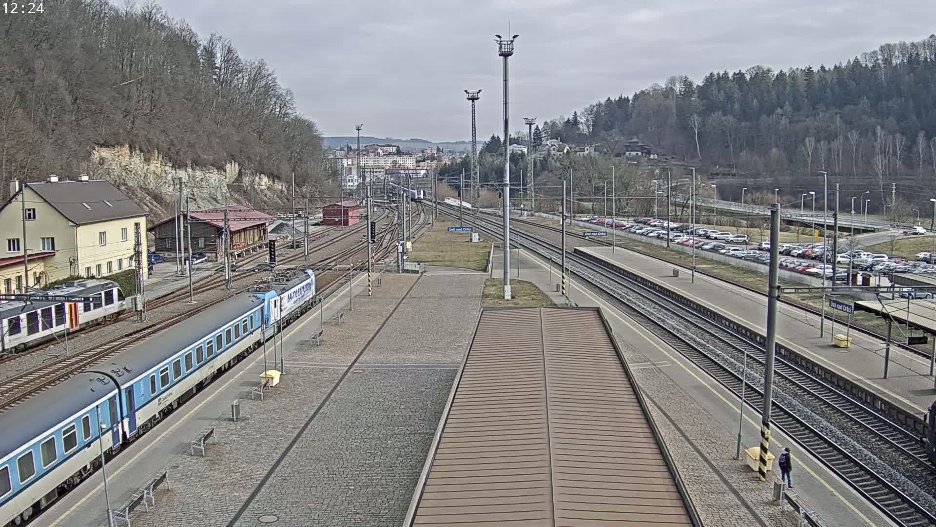 A train station with multiple train tracks, a partially visible train, and a large parking lot, all under an overcast sky.