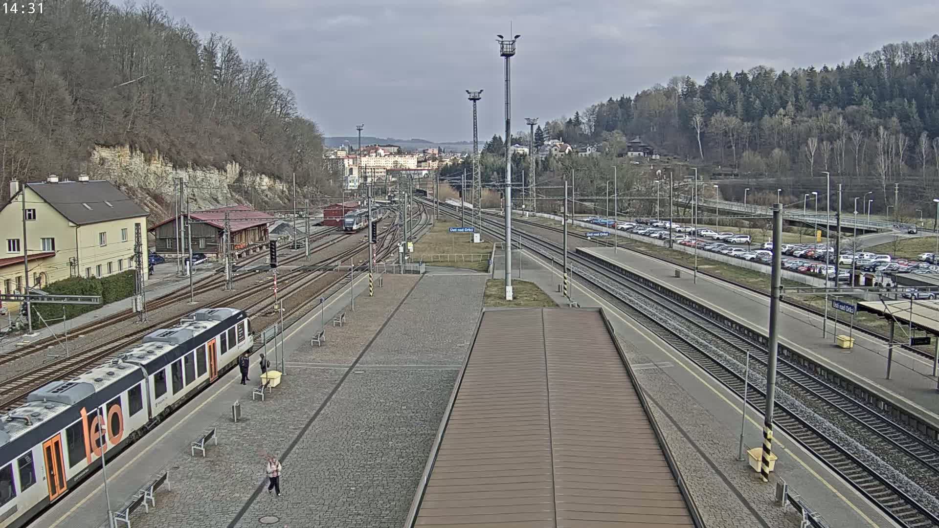 A train sits at a station platform next to multiple train tracks, with a large parking lot across the tracks and a hillside covered in leafless trees in the background under a cloudy sky.