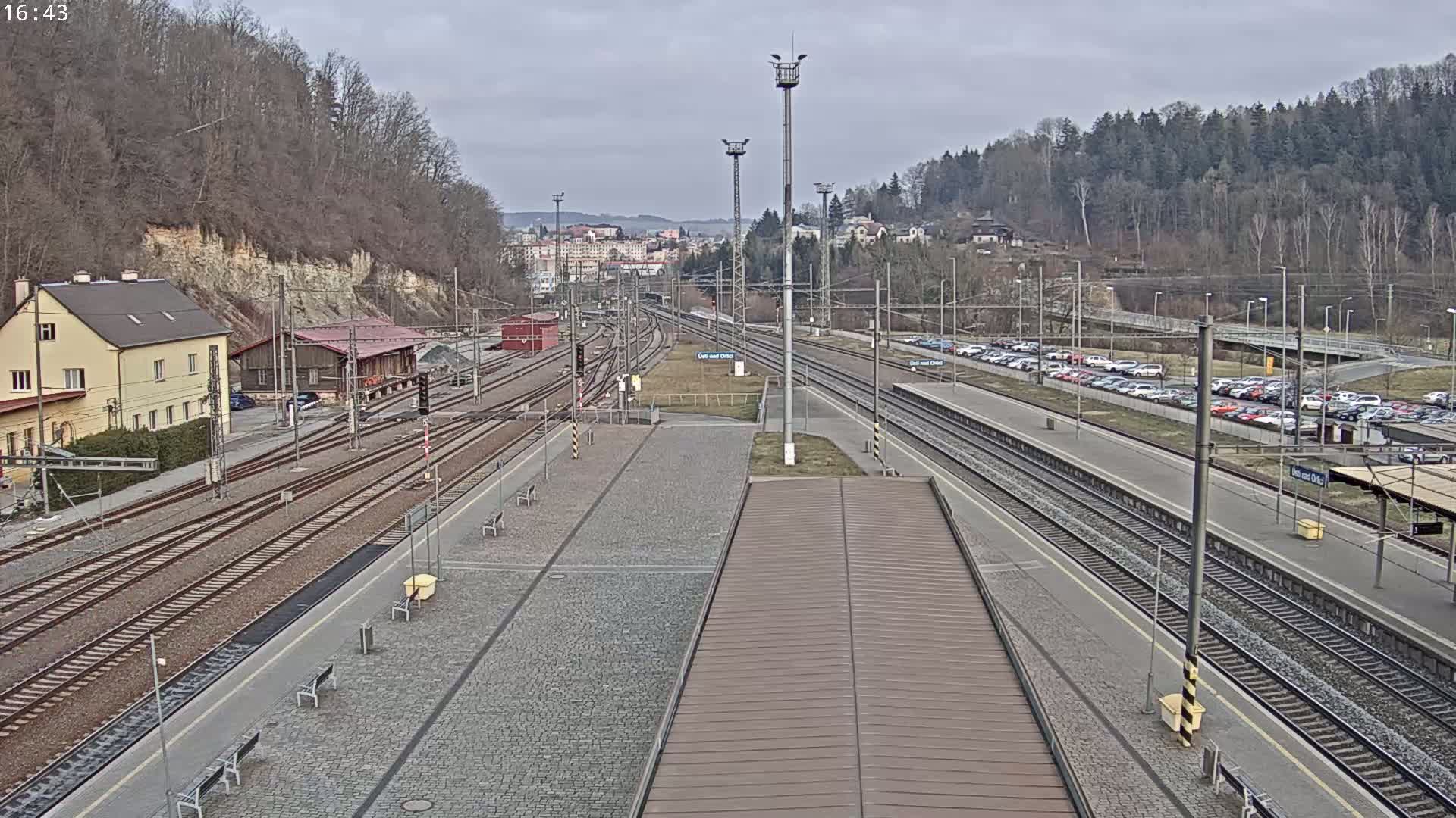 A train station with multiple tracks, a parking lot full of cars, and buildings nestled beside a tree-covered hillside on an overcast day.