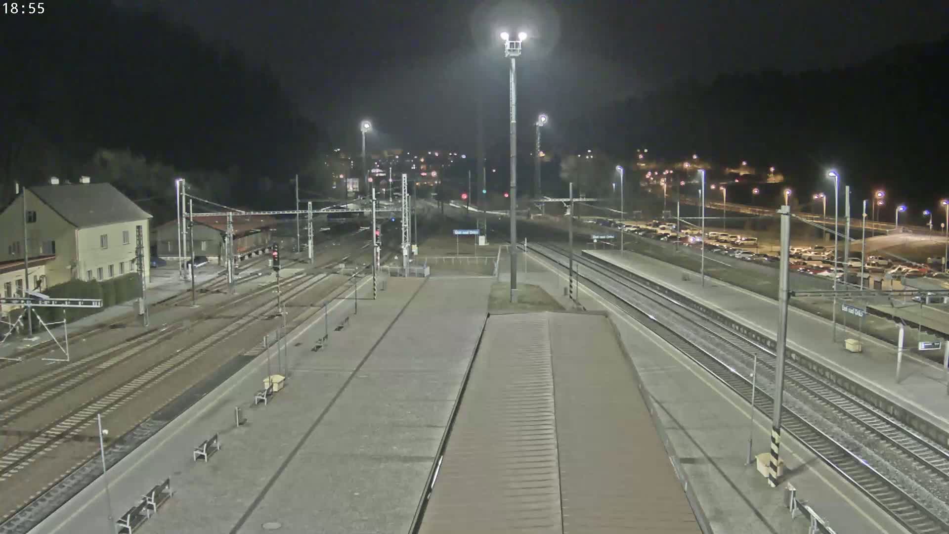 A nighttime view of a train station with multiple tracks, platforms, and a parking lot, surrounded by dark hills.
