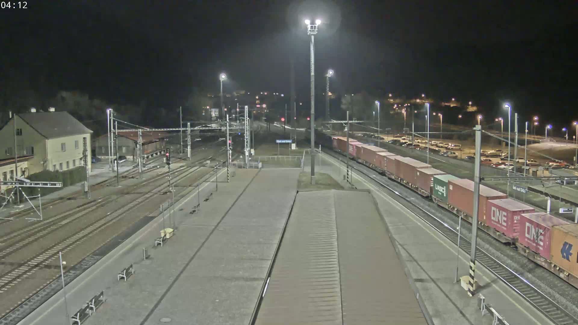A nighttime view of a train station with a freight train passing through, under bright artificial lighting.