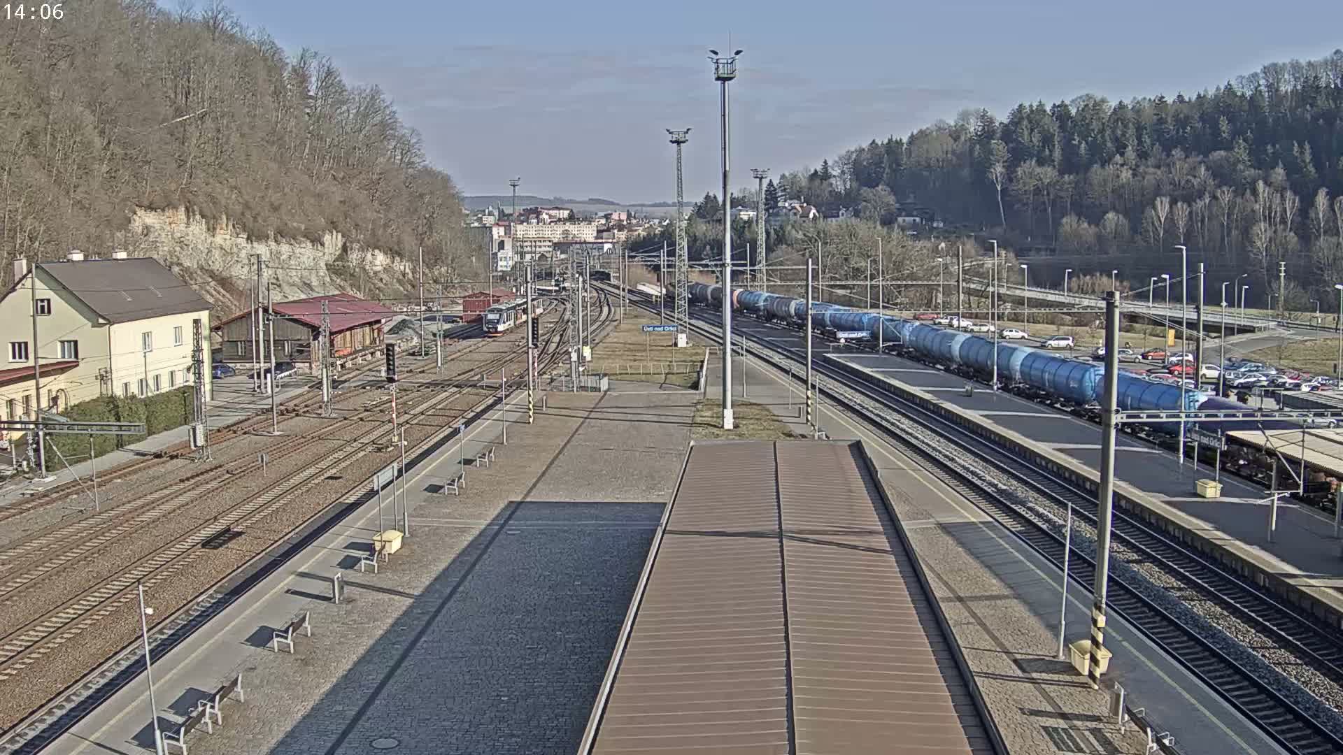 A train station on a sunny day with several train cars, including a long line of blue tanker cars, on the tracks, surrounded by buildings and trees.