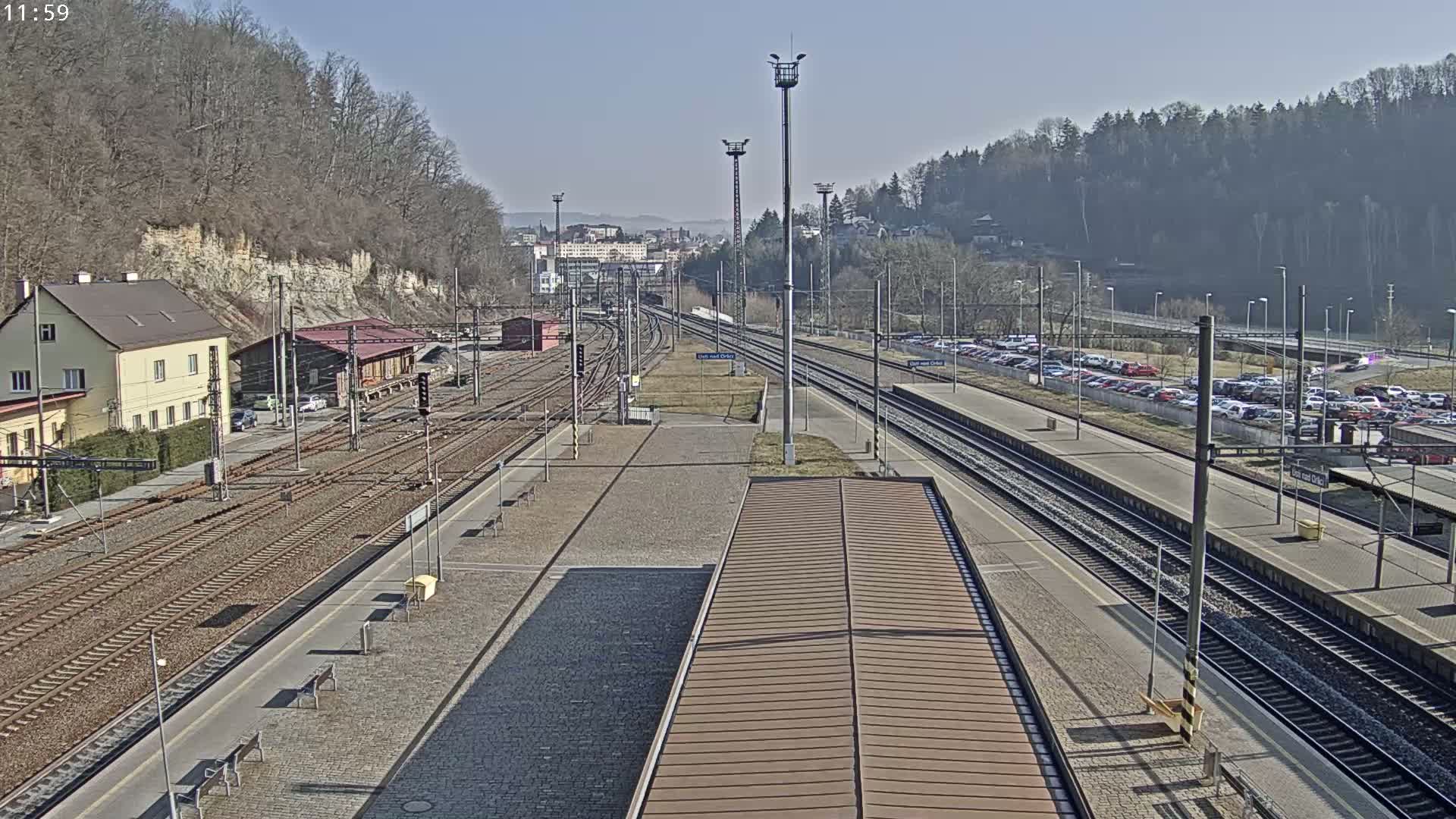 A sunny day reveals a train station with multiple tracks, platforms, and a large parking lot full of cars, all nestled beside a hillside covered in leafless trees.