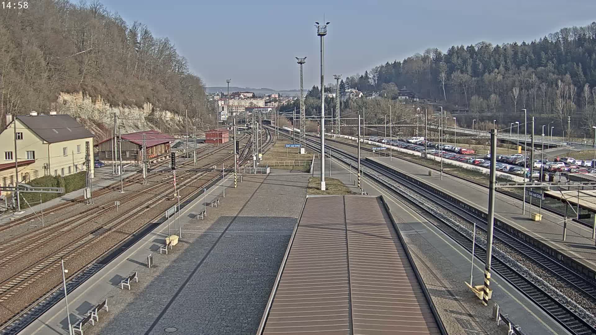A train station on a sunny day with multiple train tracks, a parking lot full of cars, and a building on one side, all surrounded by trees and a hill.