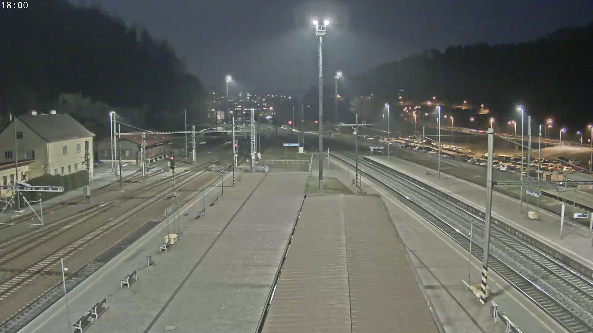 A nighttime view of a train station with multiple tracks, a platform, and a parking lot filled with cars under artificial lighting.