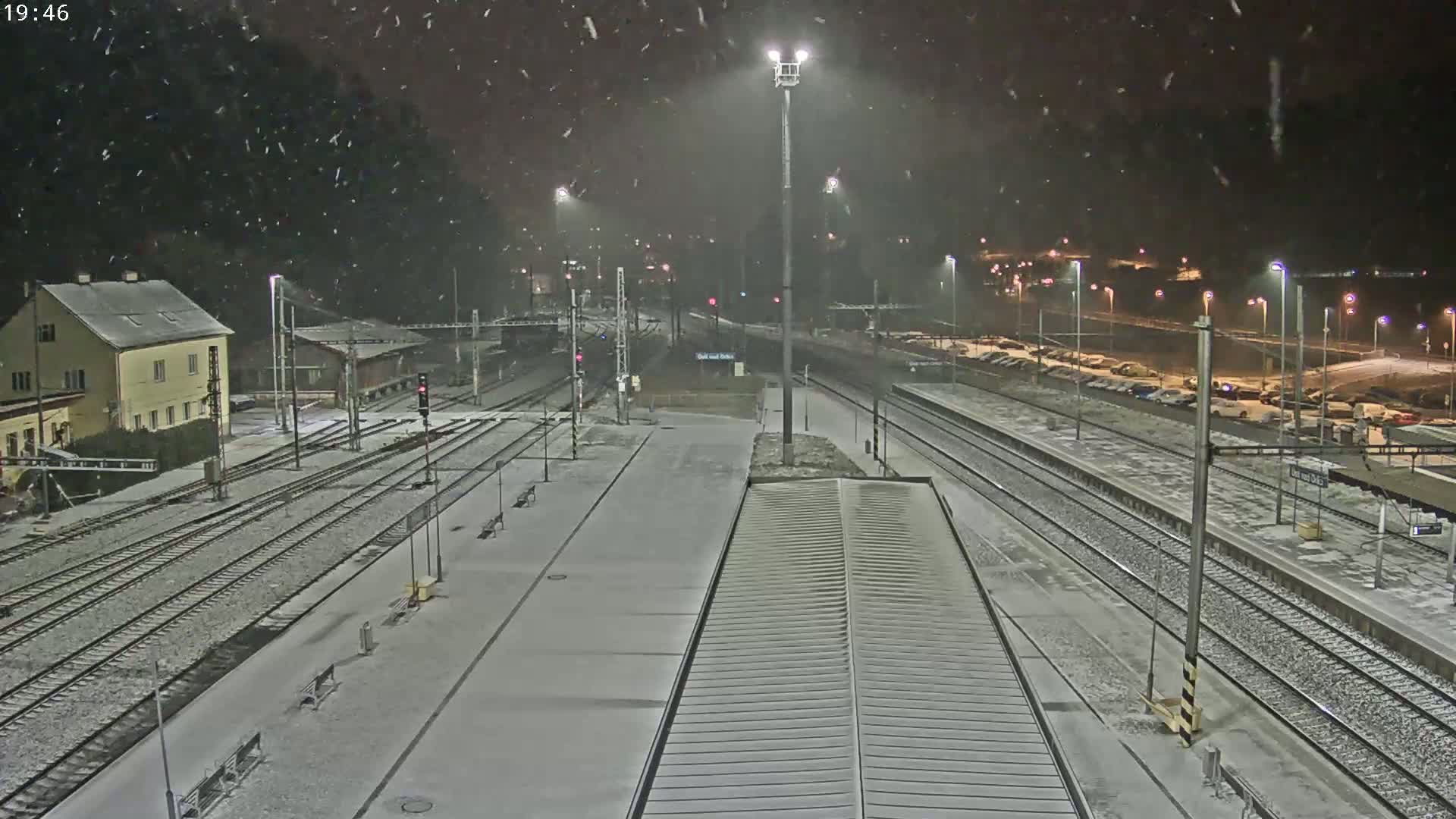 A snow-covered train station at night with multiple train tracks and a building in the background under falling snow.