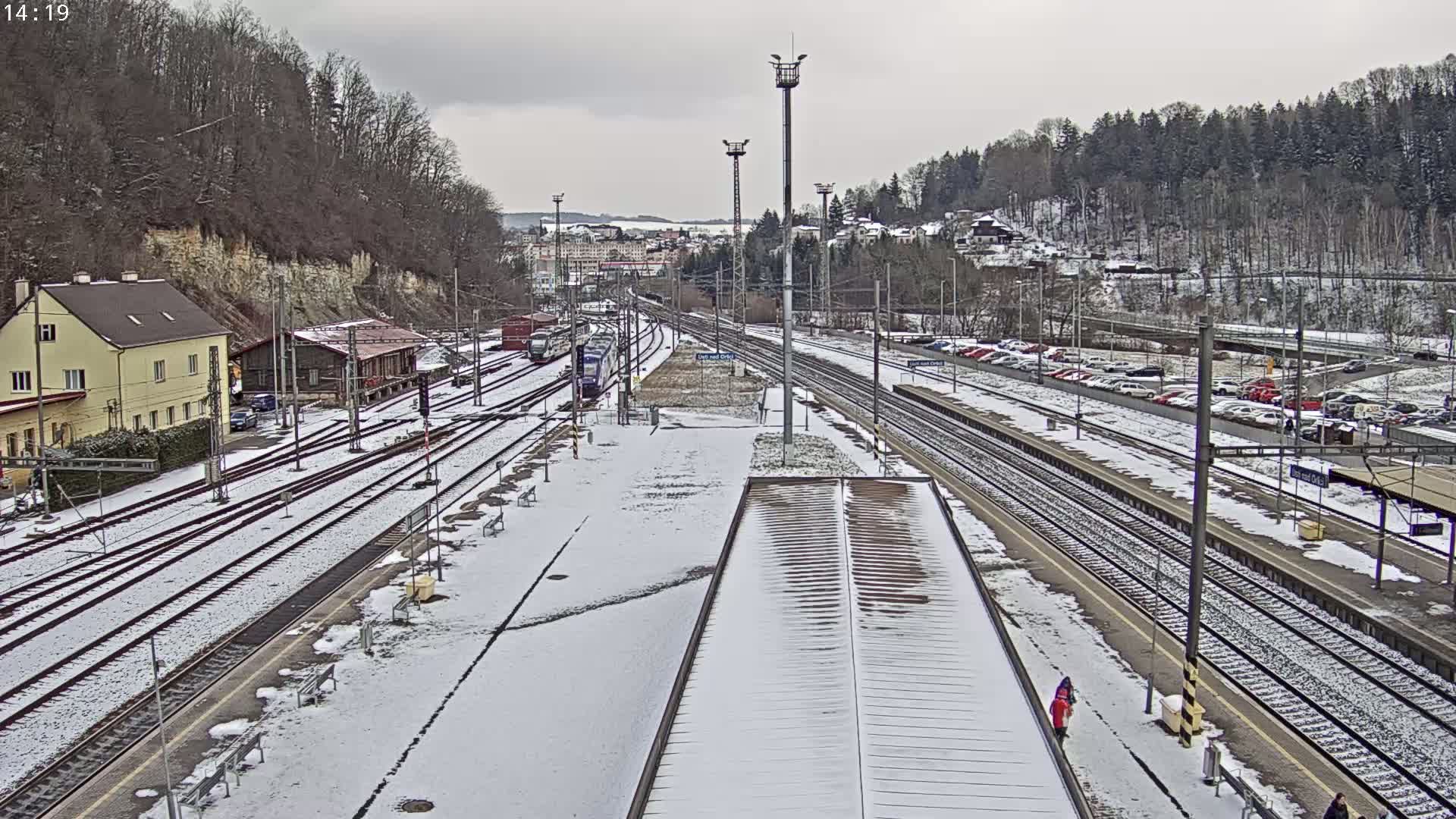 A snow-covered train station with multiple tracks and a few trains visible, surrounded by buildings and a hillside covered in leafless trees under an overcast sky.