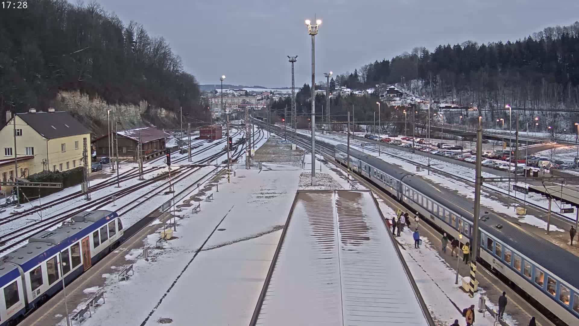 A snow-covered train station with two passenger trains and a parking lot is visible, under an overcast sky.