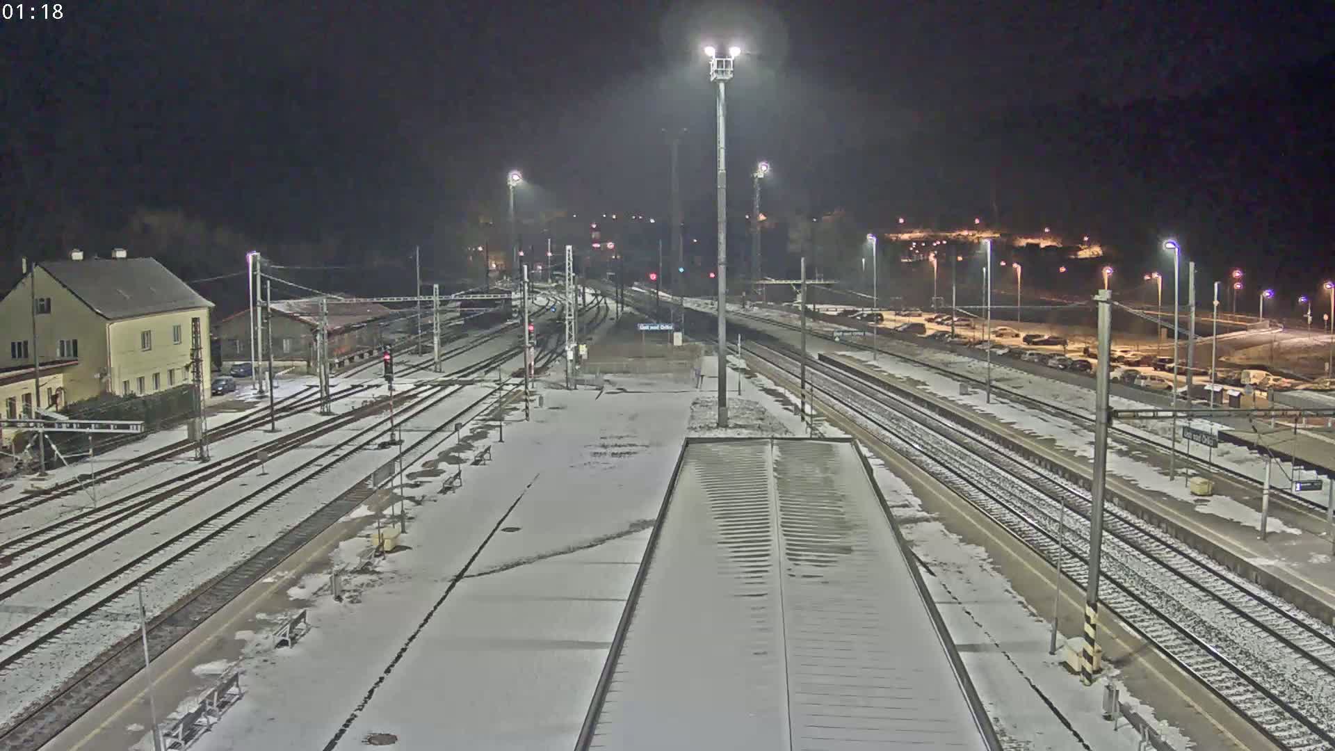 A snow-covered train station at night, illuminated by numerous lights, with multiple train tracks extending into the distance.