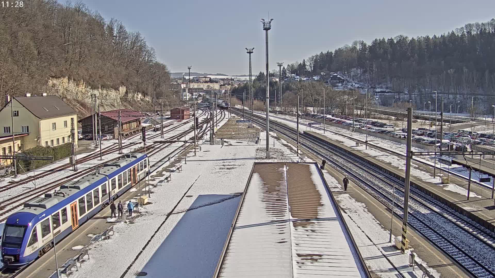 A snow-covered train station with a blue train at the platform, multiple train tracks, and a parking lot in the background under a sunny sky.