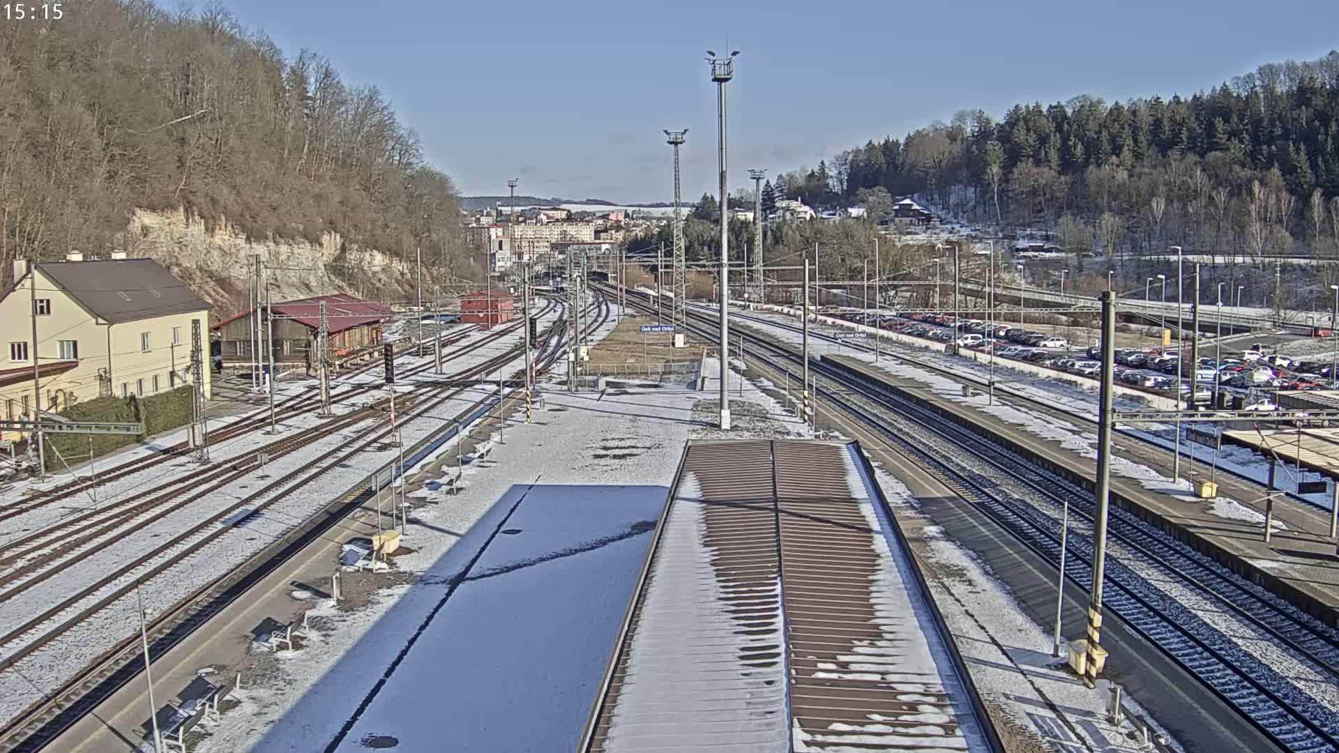 A snow-dusted train station with numerous tracks and a parking lot, situated next to a hill covered in leafless trees under a clear, sunny sky.