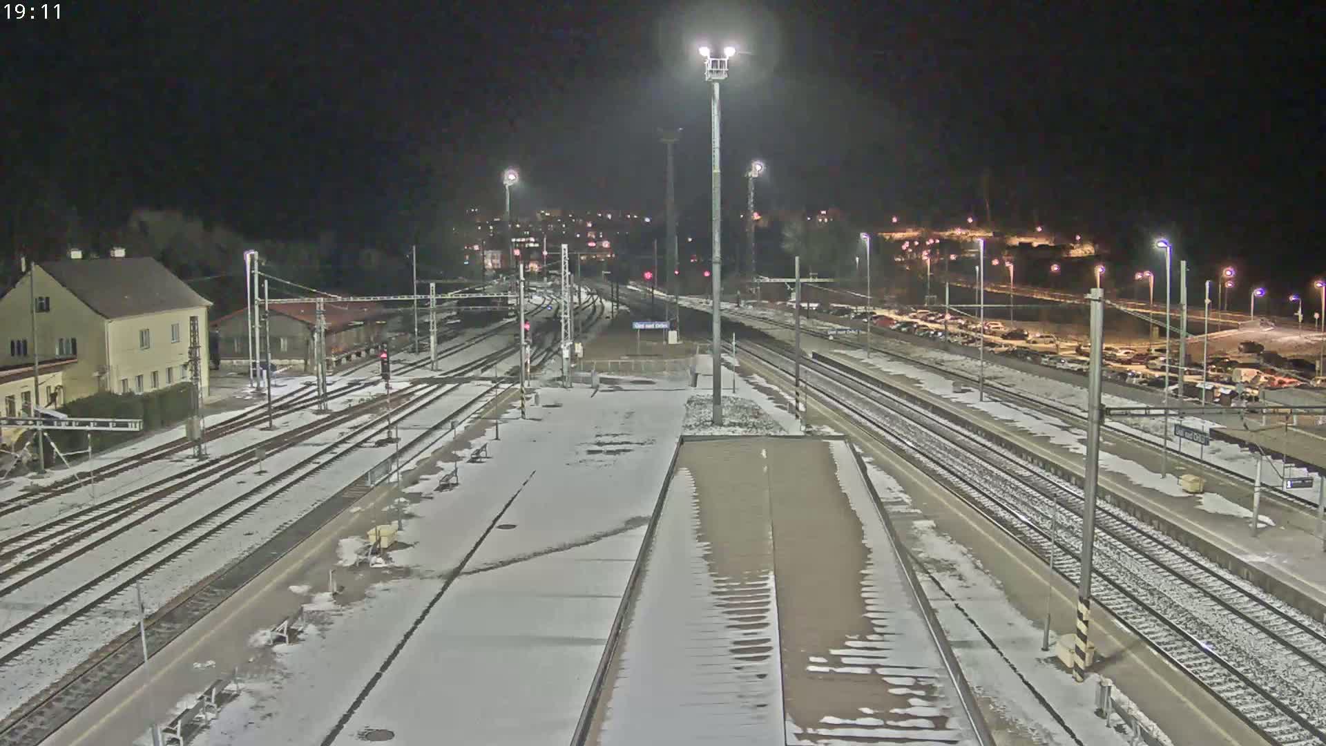 A snow-covered train station at night is illuminated by several tall lights, with multiple train tracks and a parking lot visible in the background.