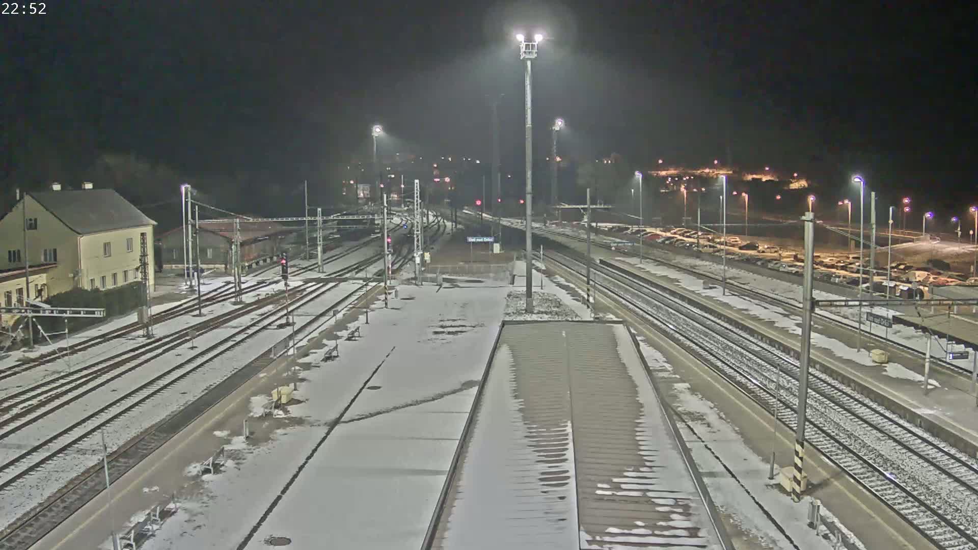 A snow-covered train station at night, illuminated by bright lights, with multiple train tracks and a parking lot visible in the background.