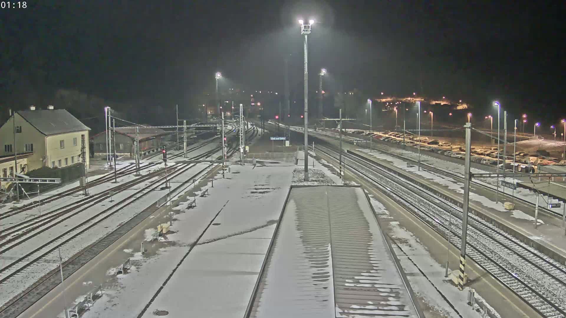 A snow-covered train station at night, illuminated by artificial lights, shows multiple train tracks and a parking lot with several cars.
