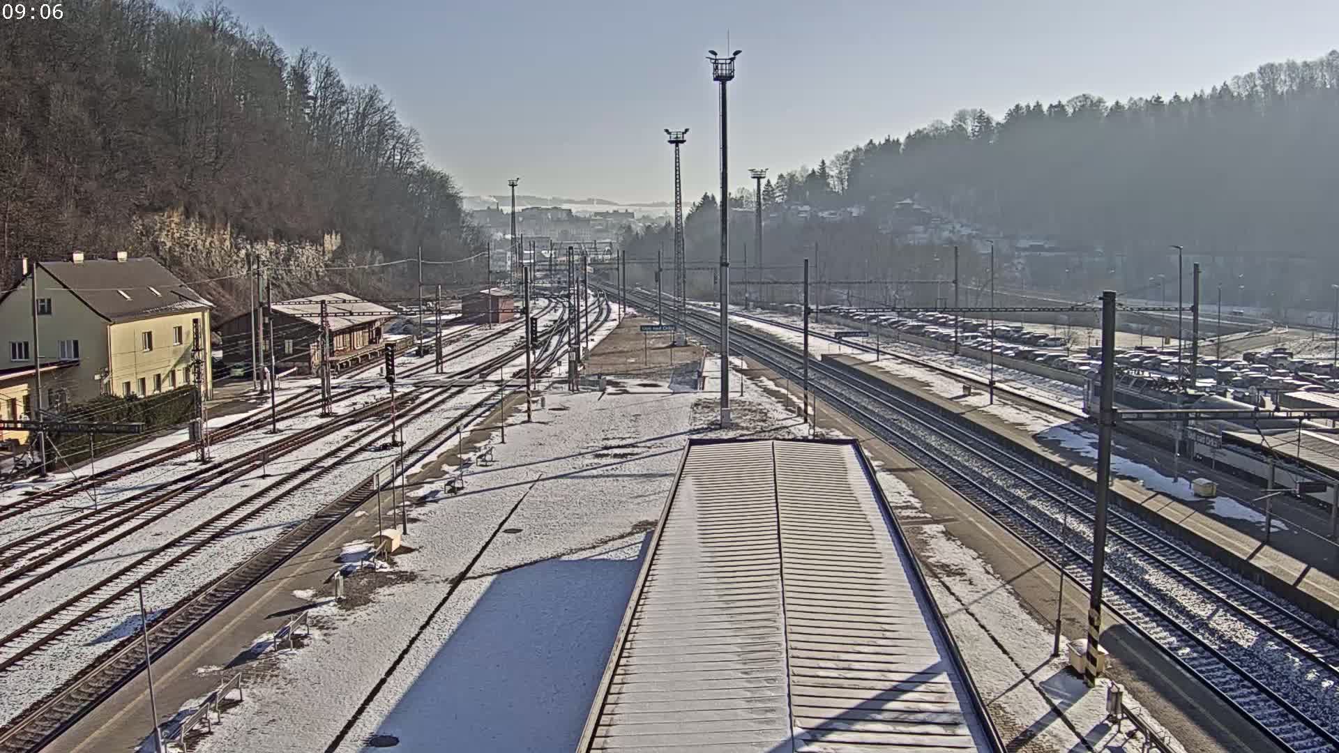 A snow-covered train station with multiple tracks, a train, and a building, situated against a backdrop of a hazy, tree-covered hillside under a clear sky.