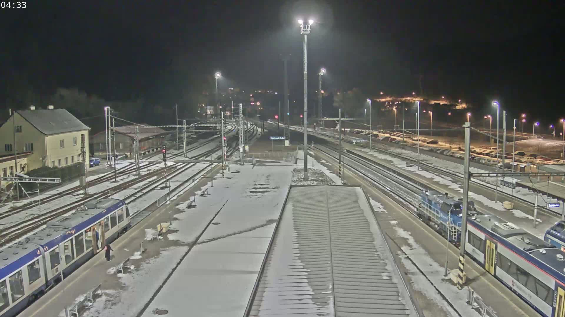 A snow-covered train station at night with several trains parked at platforms and a few cars visible in a parking lot.