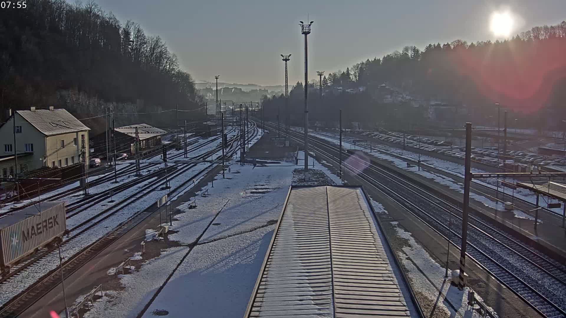 A snowy train station with multiple tracks, buildings, and a parking lot visible under a bright, sunny sky.