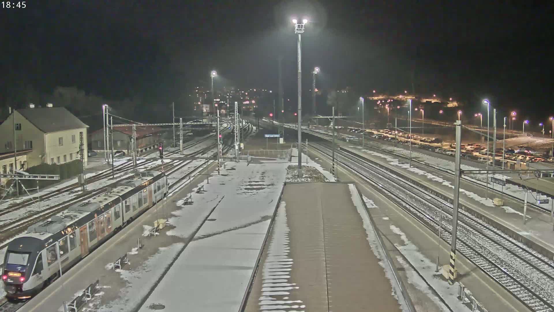 A train sits at a snow-covered nighttime train station with multiple tracks and a parking lot visible in the background.