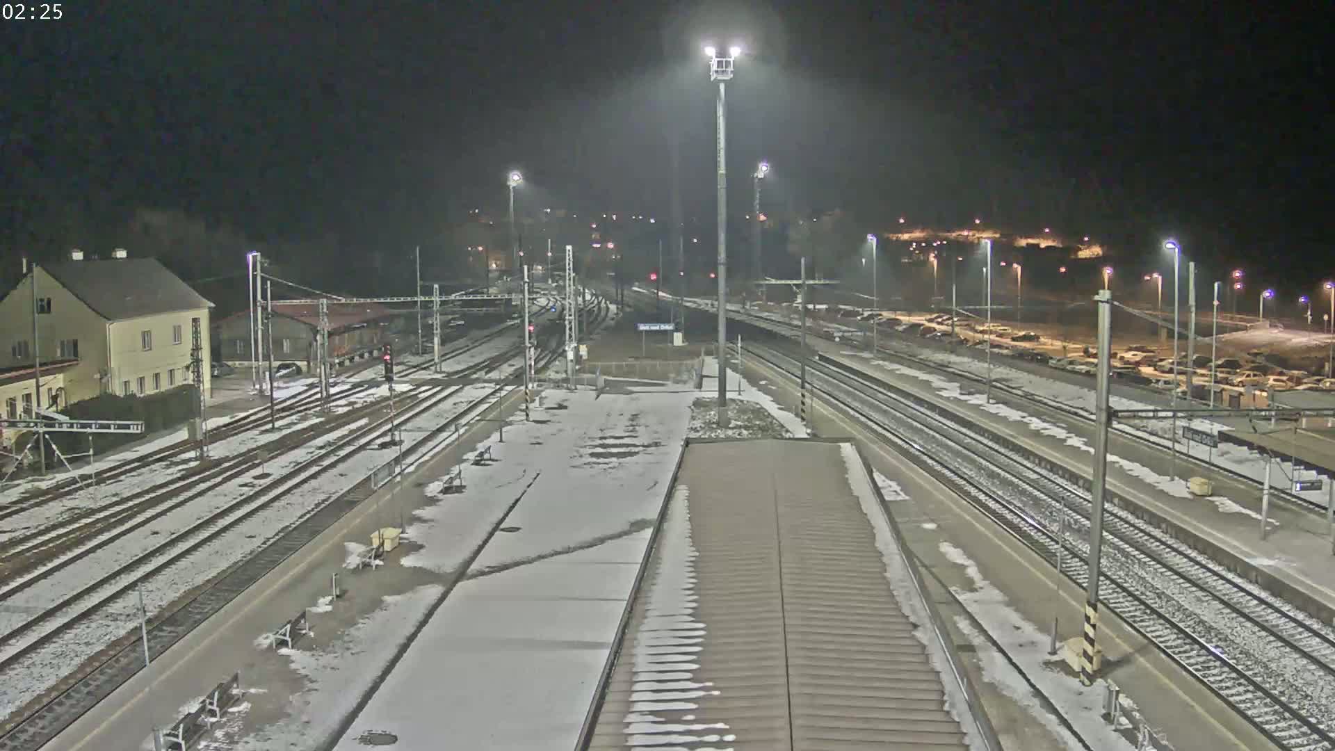 A snow-covered train station at night, with multiple train tracks and a parking lot visible.